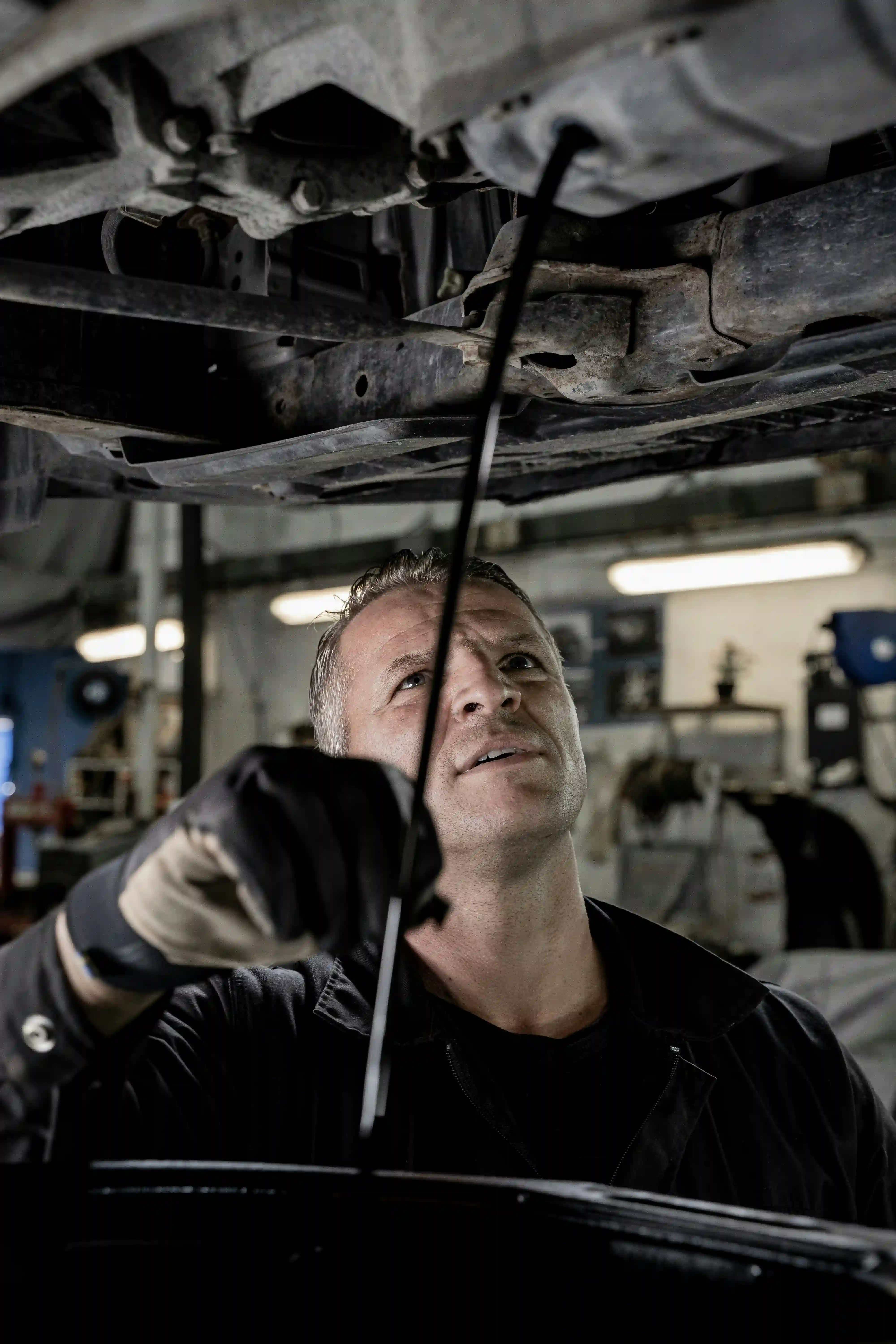 Ölwechsel in der Werkstatt: Mechaniker steht unter einem angehobenen Fahrzeug und blickt zum Unterboden, in der Hand eine dünne Sonde.