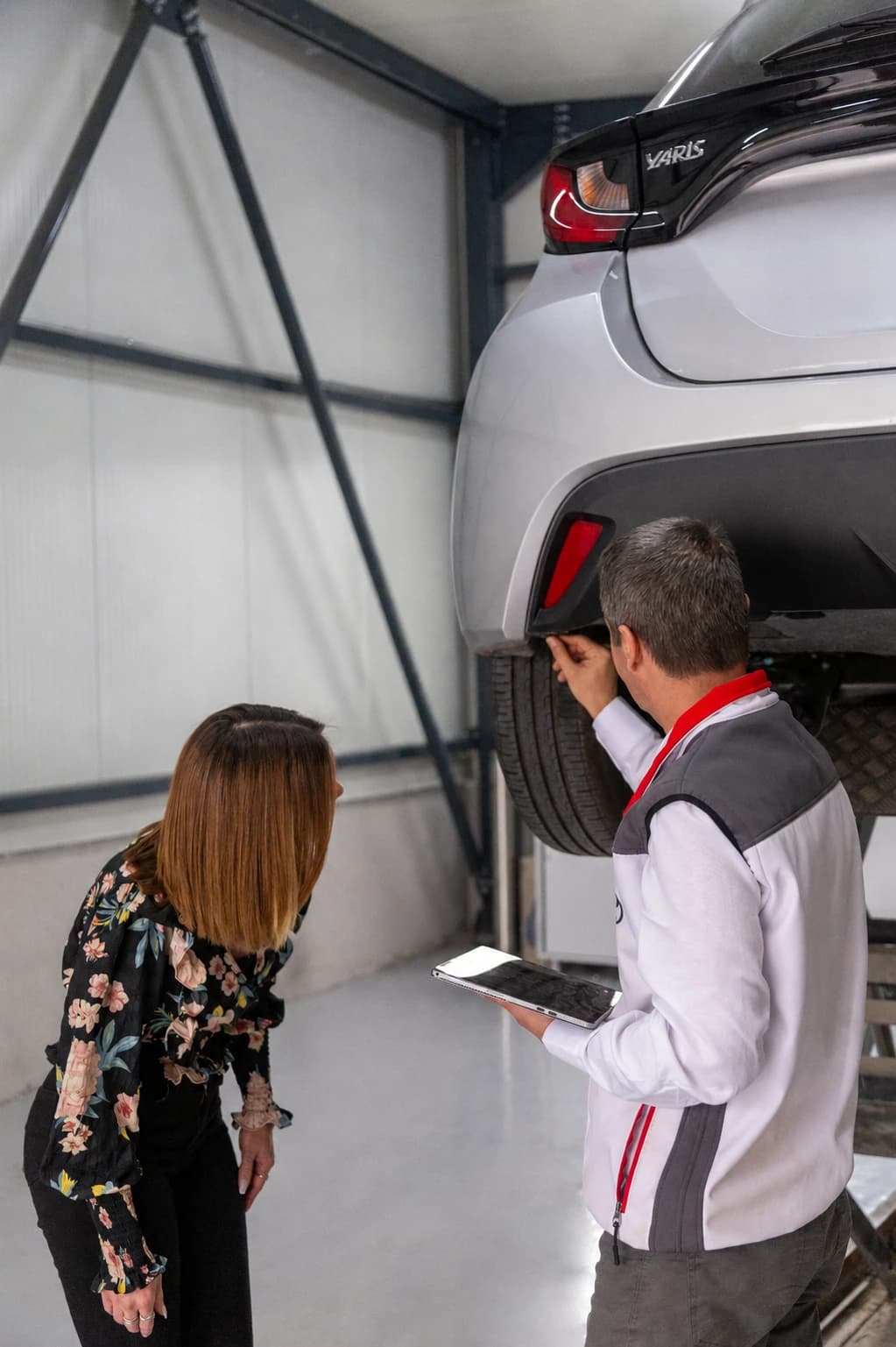 Durchsicht mit Kunden: eine Kundin und ein Mitarbeiter stehen unter einem angehobenen weißen Toyota, er zeigt mit dem Tablet in der Hand auf den Unterboden.