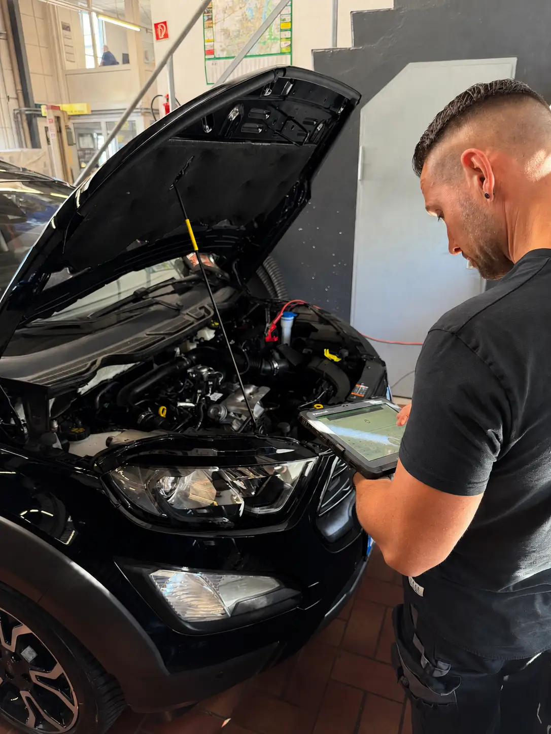 Fehlerspeicher auslesen: Mechaniker mit einem Diagnose-Tablet am geöffneten Motorraum eines schwarzen Ford.