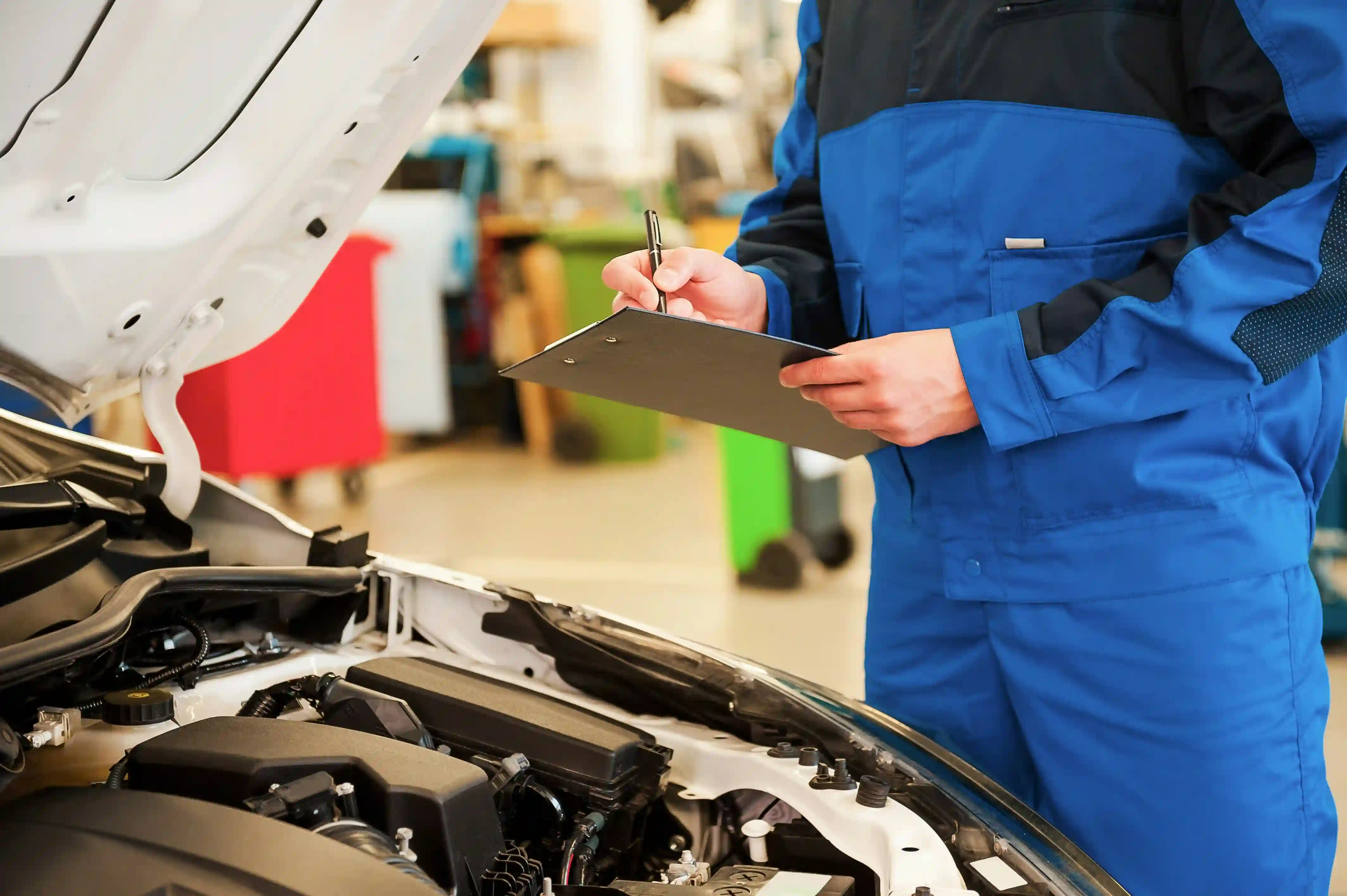 Gutachten aufnehmen: eine Person in Arbeitskleidung notiert am geöffneten Motorraum eines weißen Autos den Befund auf einem Klemmbrett.