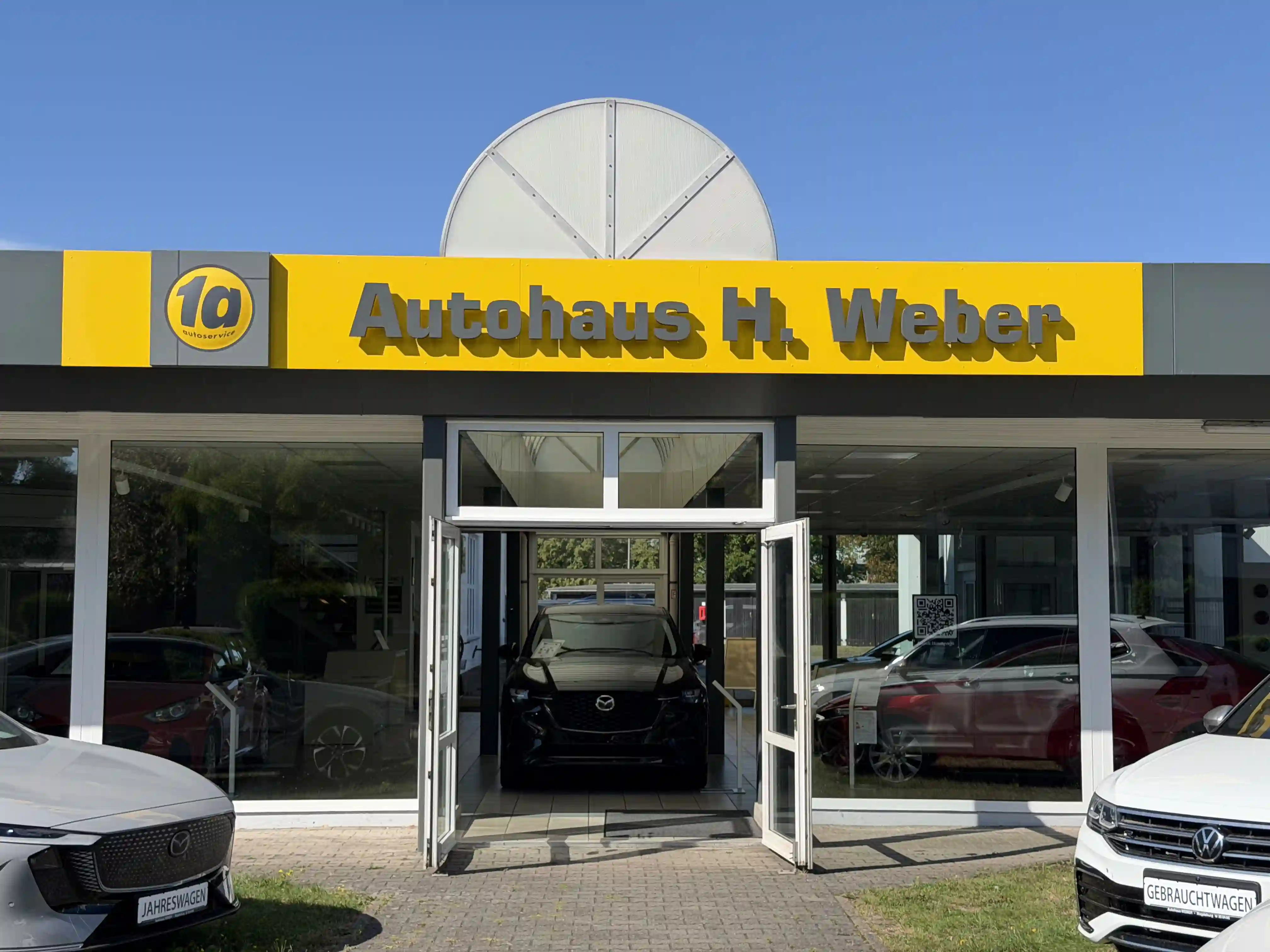 Autohaus H. Weber von der Straße aus: gelber Schriftzug über der Glasfront, offene Eingangstür, davor ein Jahreswagen und ein Gebrauchtwagen.