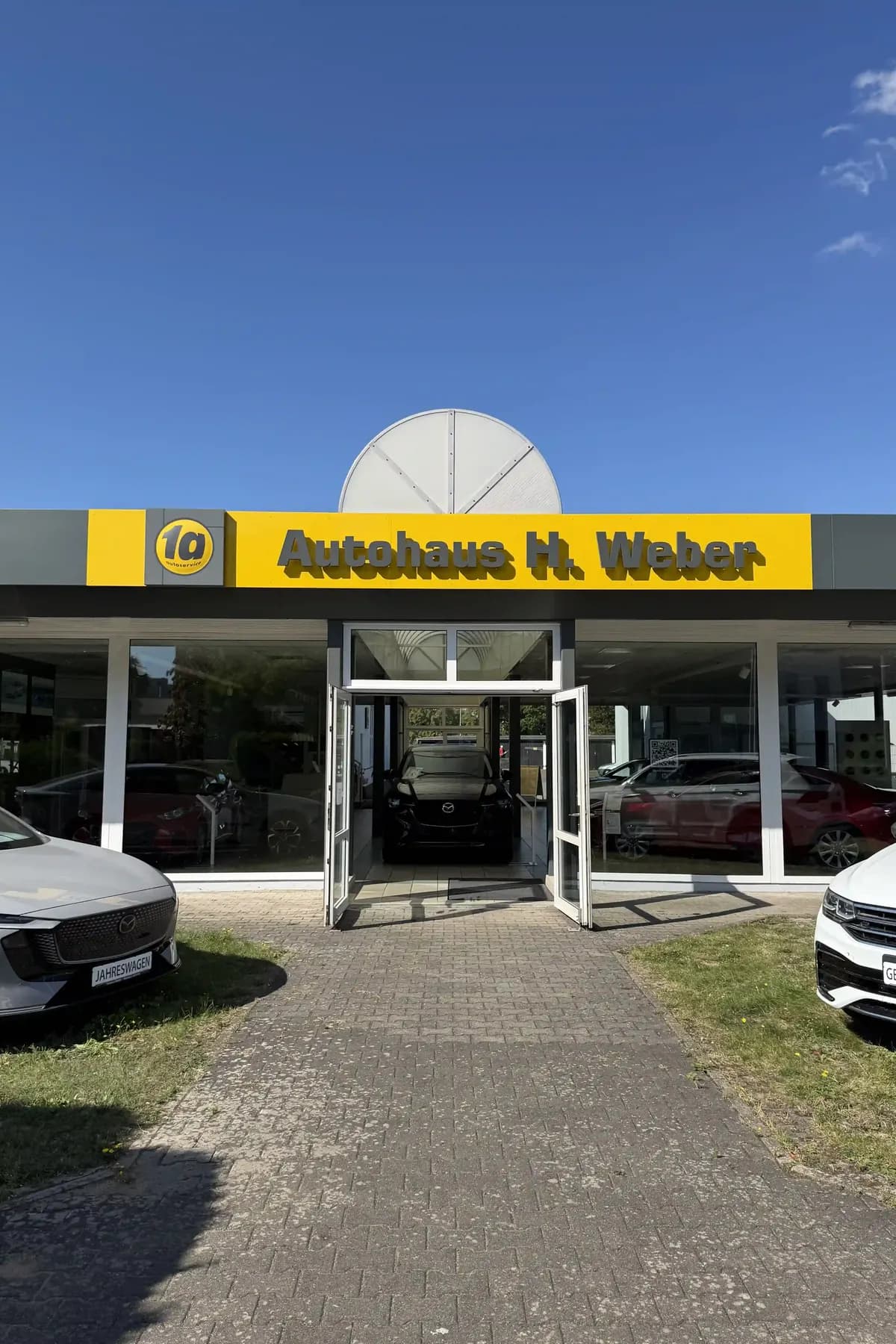 Ausstellungsraum des Autohauses mit gelbem Schriftzug, davor zwei Fahrzeuge